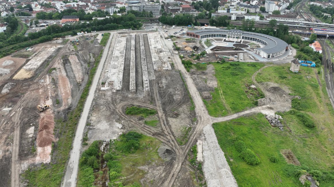 Gelände mit Grün- und Baustellenanteil. Im Hintergrund befindet sich ein halbrundes, flaches Gebäude und weitere Häuser.