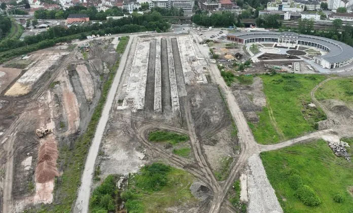 Luftaufnahme eines Geländes, auf dem Grün- und Baustellenflächen zu sehen sind. Im Hintergrund befindet sich ein flaches, halbrundes Gebäude und weitere Häuser.