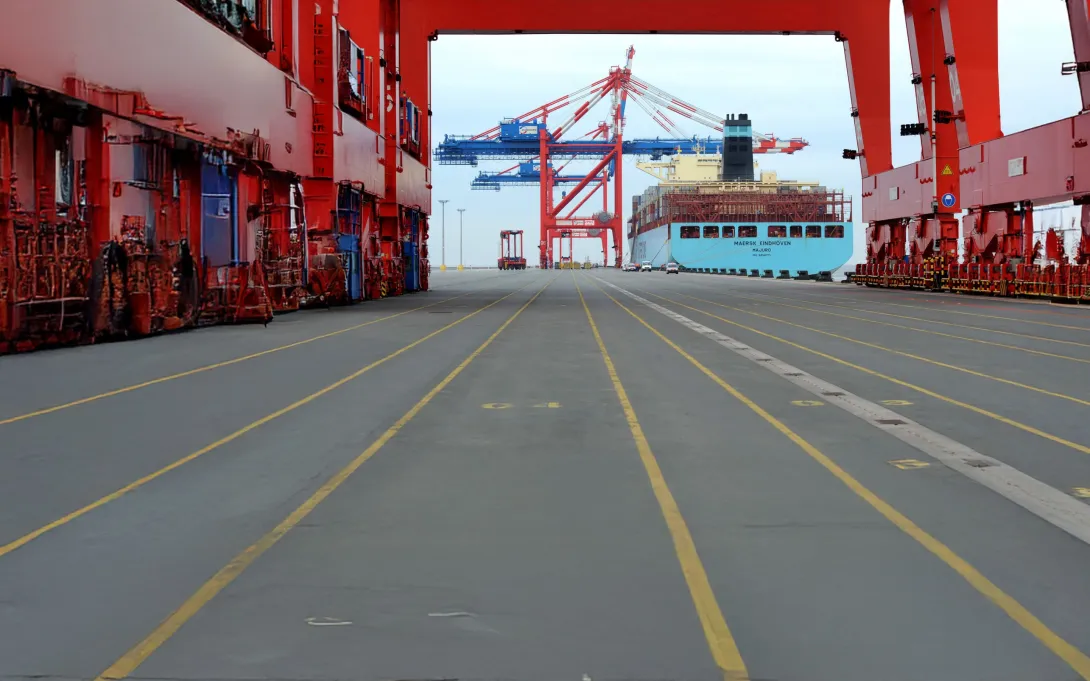 Frachthafen, der von einem roten, stählernen Konstrukt überspannt wird. Im Hintergrund liegt ein hellblaues Frachtschiff unter großen Kränen an.