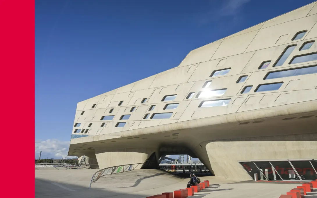 Futuristisches Betongebäude mit unregelmässigen Fensterformen und grossen Überhängen vor blauem Himmel, im Vordergrund rote Sitzblöcke und eine Rampe.