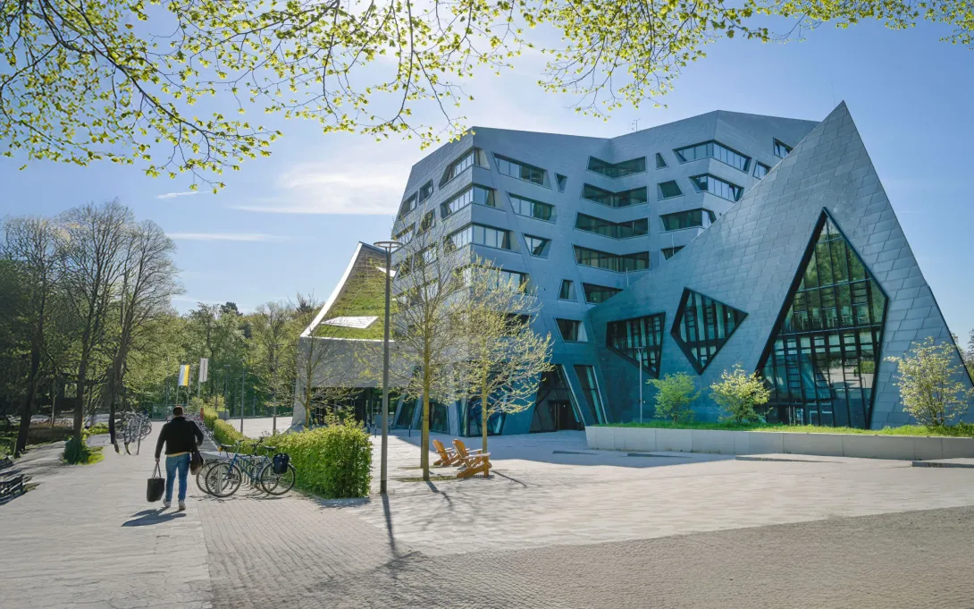 Modernes Gebäude mit auffälliger, kantiger Architektur und grossen Glasflächen, umgeben von Bäumen und Fahrrädern, bei sonnigem Frühlingswetter.