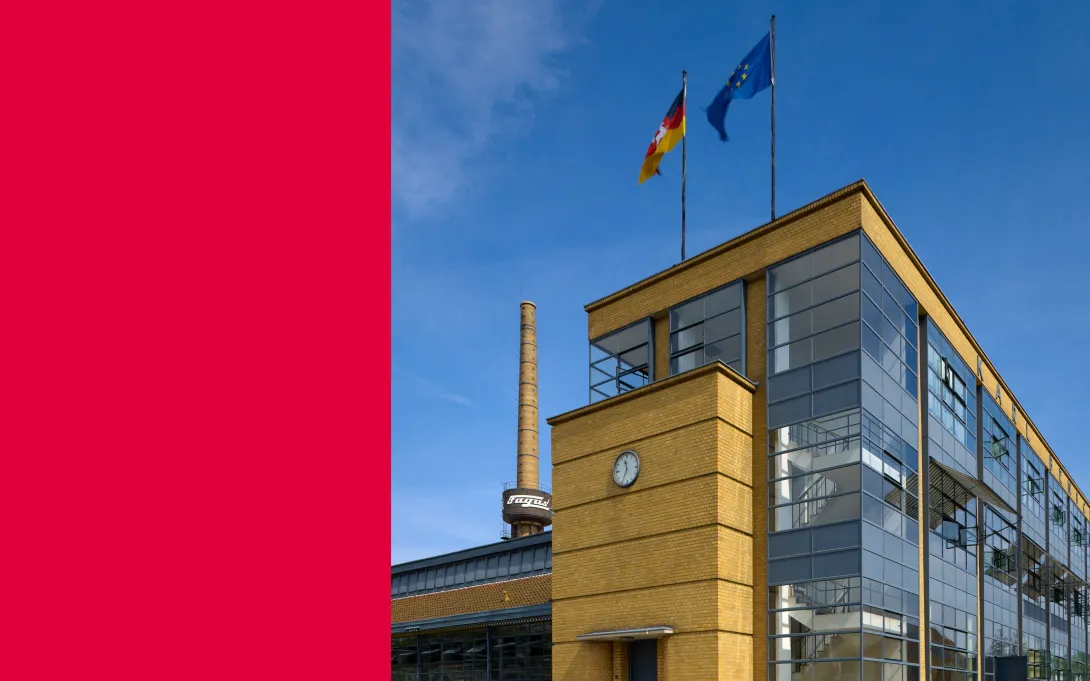 Gelbes Gebäude mit großen Glasflächen vor blauem Himmel. Auf dem Dach wehen eine Deutschland- und eine Europafahne.